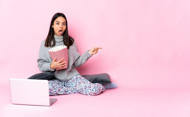 Young mixed race woman eating popcorn while watching a movie on the laptop with surprise expression while pointing side