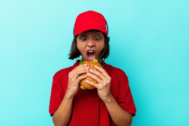 Young mixed race woman fast food restaurant worker holding a burger isolated on blue background