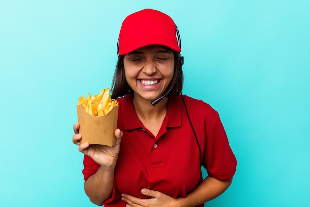 젊은 혼혈 여성 패스트푸드 레스토랑 직원이 파란 배경에 격리된 감자튀김을 들고 웃고 즐겁게 지내고 있습니다.