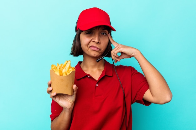 若い混血女性ファーストフード レストランの従業員は、青い背景に分離されたフライド ポテトを持ち、寺院を指で指し、考え、仕事に集中しました。