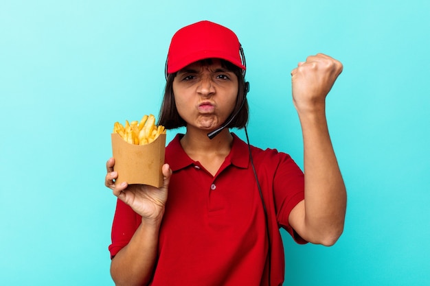 青の背景にフライド ポテトを保持している若い混血女性ファーストフード レストランの労働者は、カメラに拳を見せ、積極的な表情を見せています。