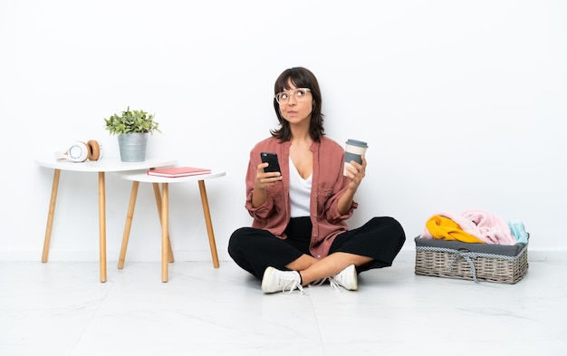 Young mixed race woman folding clothes sitting on the floor isolated on white background holding coffee to take away and a mobile while thinking something