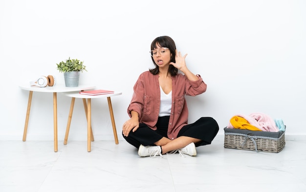 Young mixed race woman folding clothes sitting on the floor isolated on white background listening to something by putting hand on the ear