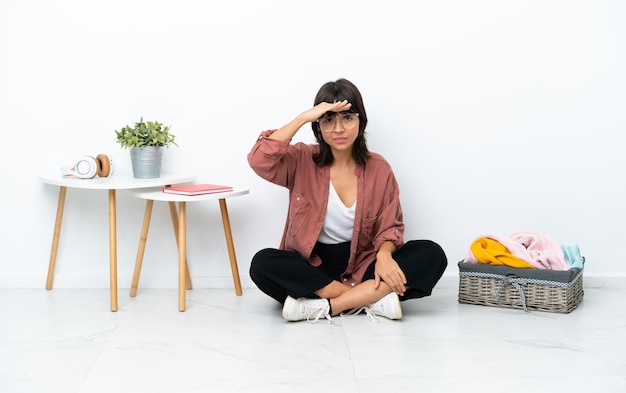 Young mixed race woman folding clothes sitting on the floor isolated on white background looking far away with hand to look something