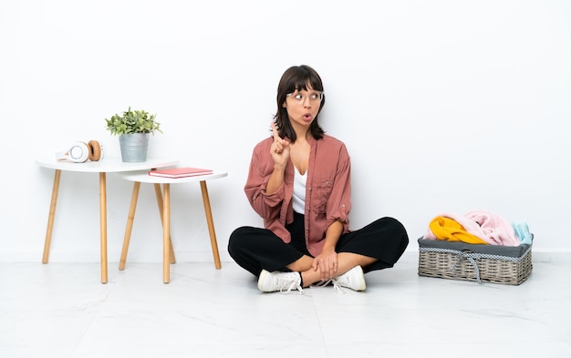 Young mixed race woman folding clothes sitting on the floor isolated on white background thinking an idea pointing the finger up
