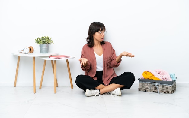 Young mixed race woman folding clothes sitting on the floor isolated on white background with surprise facial expression