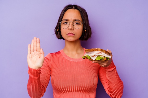 사진 젊은 혼합 된 경주 여자 당신을 방지하는 정지 신호를 보여주는 뻗은 손으로 서 보라색 배경에 고립 된 샌드위치를 들고.