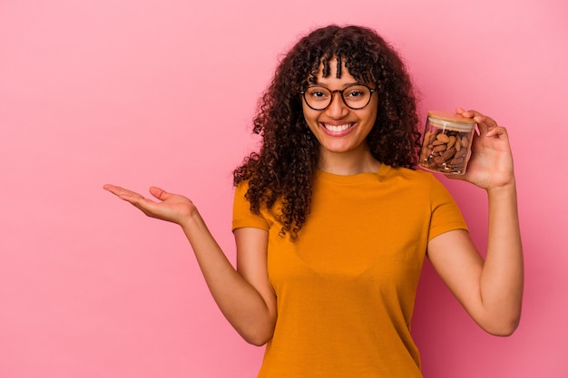 Young mixed race woman holding an almond jar isolated on pink background showing a copy space on a palm and holding another hand on waist.