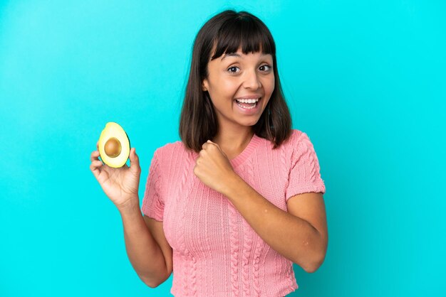 Young mixed race woman holding an avocado isolated on blue background celebrating a victory