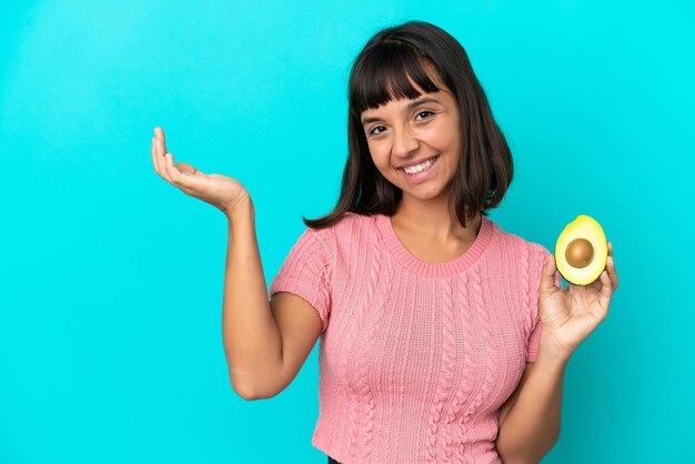 青い背景に分離されたアボカドを持っている若い混血の女性は、来て招待するために手を横に伸ばします