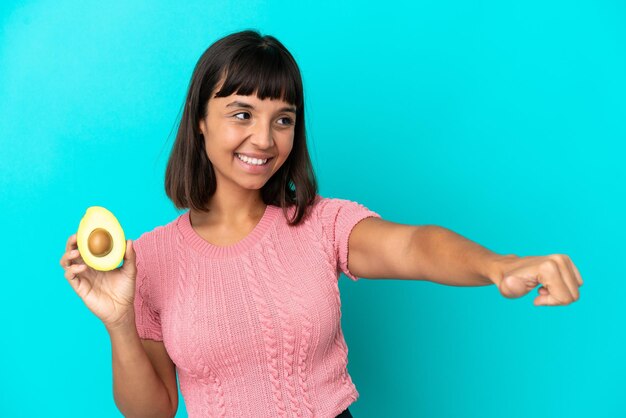 親指を立てるジェスチャーを与える青い背景で隔離のアボカドを保持している若い混血の女性