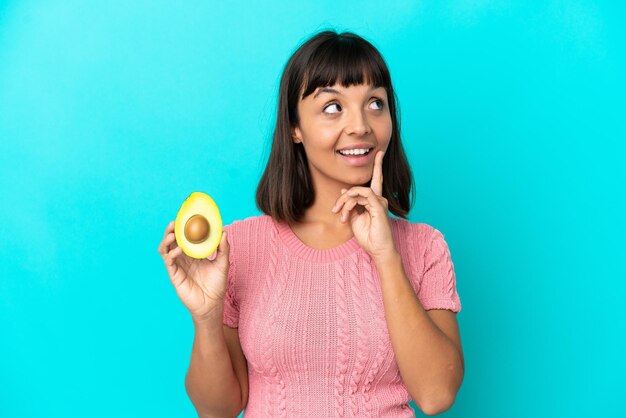 Young mixed race woman holding an avocado isolated on blue background thinking an idea while looking up