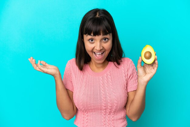 Giovane donna di razza mista in possesso di un avocado isolato su sfondo blu con espressione facciale scioccata
