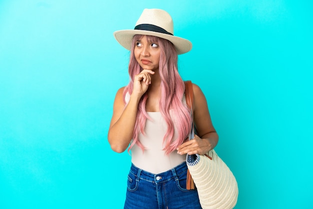 Young mixed race woman holding a beach bag with pamela isolated on blue background having doubts and with confuse face expression