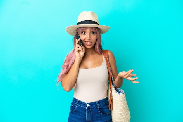 Young mixed race woman holding a beach bag with pamela isolated on blue background keeping a conversation with the mobile phone with someone