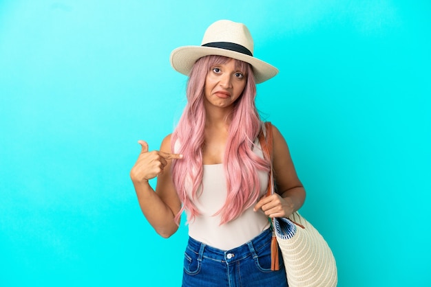 Young mixed race woman holding a beach bag with pamela isolated on blue background proud and self-satisfied