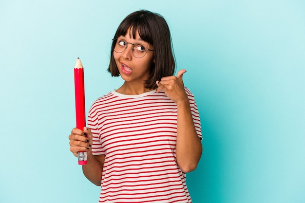 Young mixed race woman holding a big pencil isolated on blue background showing a mobile phone call gesture with fingers.