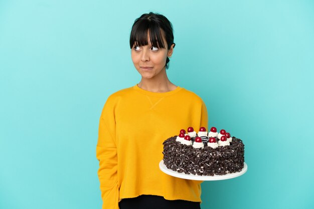Молодая женщина смешанной расы держит торт ко дню рождения, делая жест сомнения, глядя в сторону