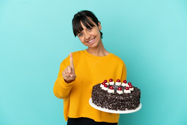 Молодая женщина смешанной расы держит торт ко дню рождения, показывая и поднимая палец
