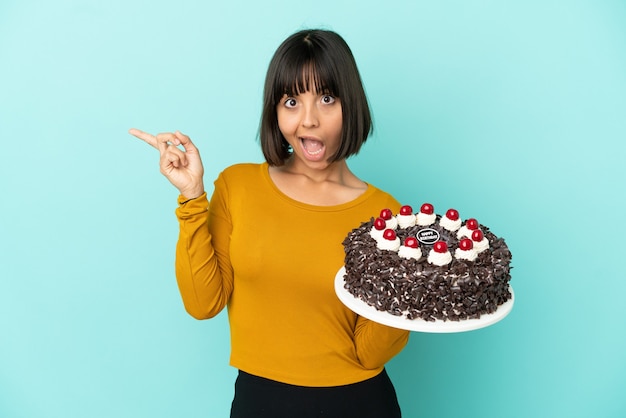 Young mixed race woman holding birthday cake surprised and pointing finger to the side