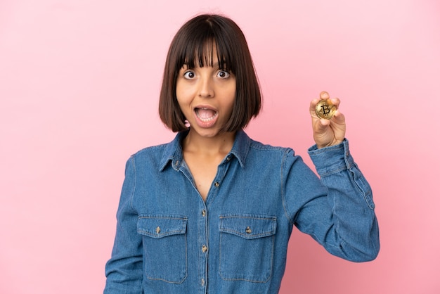 Young mixed race woman holding a Bitcoin isolated background with surprise facial expression