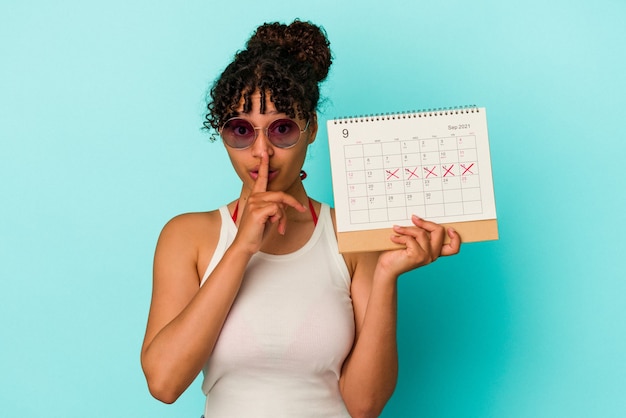 Foto giovane donna di razza mista che tiene il calendario isolato su sfondo blu mantenendo un segreto o chiedendo silenzio.