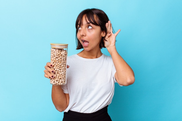 Young mixed race woman holding chickpeas jar isolated on white background trying to listening a gossip.