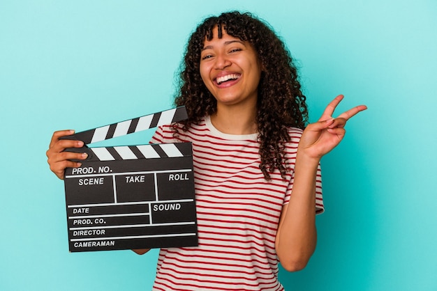 Young mixed race woman holding a clapperboard isolated on blue background joyful and carefree showing a peace symbol with fingers.