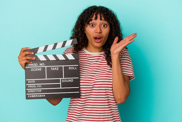 Young mixed race woman holding a clapperboard isolated on blue background surprised and shocked.