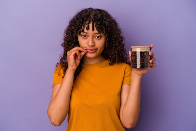 Young mixed race woman holding a coffee beans bottle isolated on purple background with fingers on lips keeping a secret.