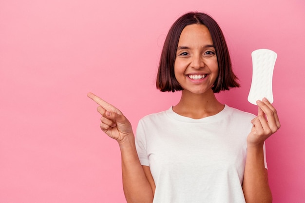 Young mixed race woman holding a compress isolated on pink background smiling and pointing aside, showing something at blank space.