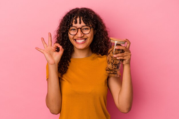 ピンクの背景に分離されたクッキーの瓶を持っている若い混血の女性は、陽気で自信を持って大丈夫なジェスチャーを示しています。