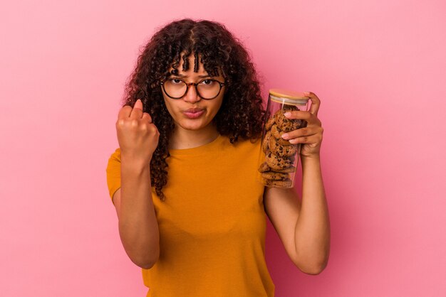 Young mixed race woman holding a cookies jar isolated on pink background showing fist to camera, aggressive facial expression.