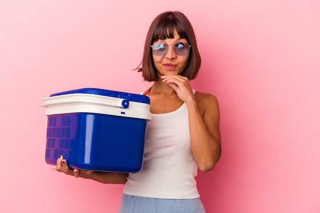 Young mixed race woman holding a cooler isolated on pink background looking sideways with doubtful and skeptical expression.