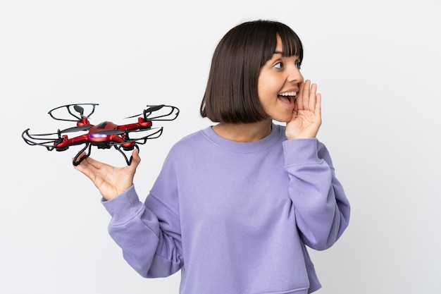 Young mixed race woman holding a drone isolated on white background shouting with mouth wide open to the side