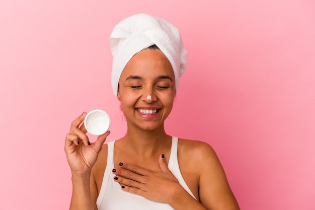 Young mixed race woman holding facial cream isolated on pink background laughs out loudly keeping hand on chest.