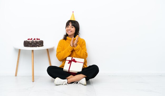 Photo young mixed race woman holding a gift sitting on the floor isolated on white background applauding after presentation in a conference