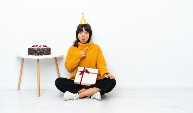 Young mixed race woman holding a gift sitting on the floor\
isolated on white background surprised and pointing side