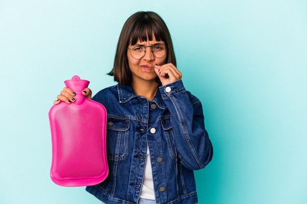 Young mixed race woman holding a hot bottle water isolated on blue background with fingers on lips keeping a secret.