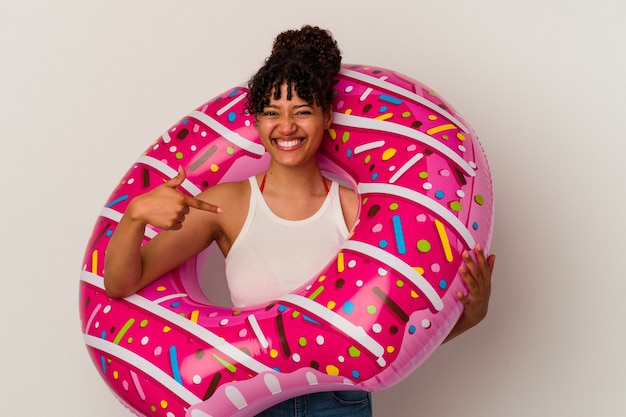 Young mixed race woman holding an inflatable air donut isolated on white wall person pointing by hand to a shirt copy space, proud and confident