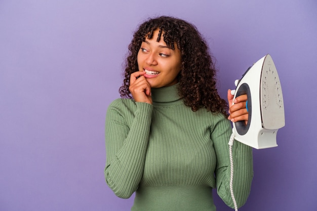 Young mixed race woman holding an iron isolated on purple background relaxed thinking about something looking at a copy space.