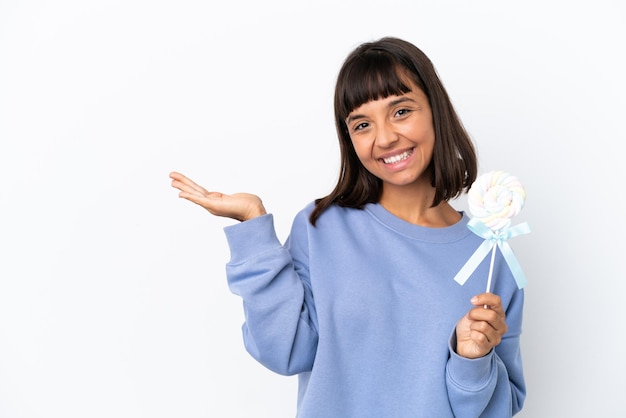 Giovane donna di razza mista che tiene in mano un lecca-lecca isolato su sfondo bianco che allunga le mani di lato per invitare a venire