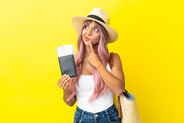 Young mixed race woman holding passport and beach bag isolated on yellow background having doubts while looking up