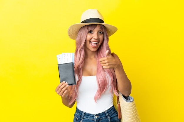 Young mixed race woman holding passport and beach bag isolated on yellow background with thumbs up because something good has happened