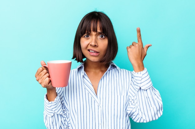 Young mixed race woman holding a pink mug isolated on blue background showing a disappointment gesture with forefinger.