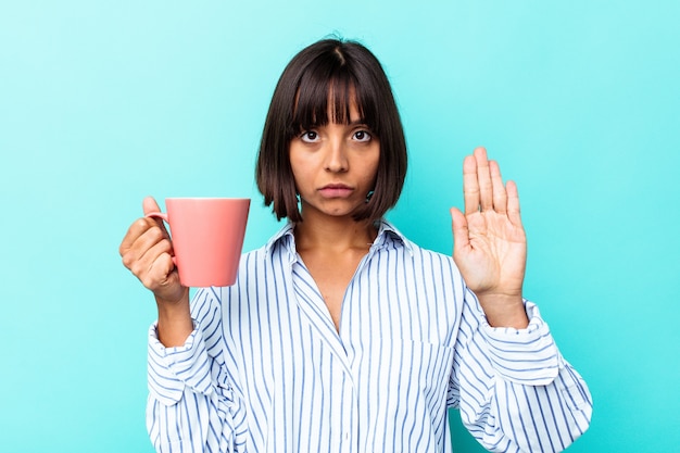 Young mixed race woman holding a pink mug isolated on blue background standing with outstretched hand showing stop sign, preventing you.