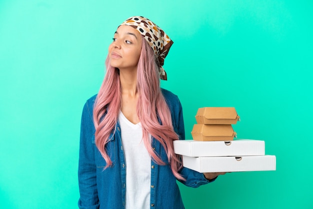 Young mixed race woman holding pizzas and burgers isolated on green background looking to the side and smiling