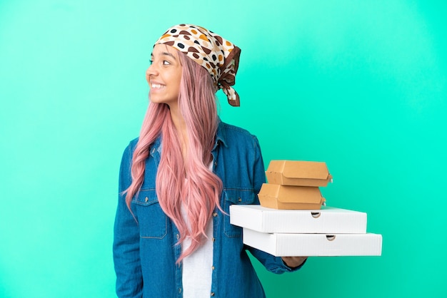 Молодая женщина смешанной расы, держащая пиццу и гамбургеры, изолированные на зеленом фоне, смотрящую сторону