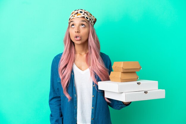 Young mixed race woman holding pizzas and burgers isolated on green background looking up and with surprised expression