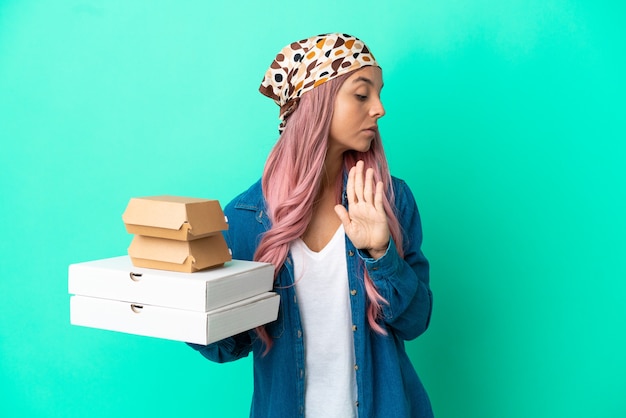 Young mixed race woman holding pizzas and burgers isolated on green background making stop gesture and disappointed